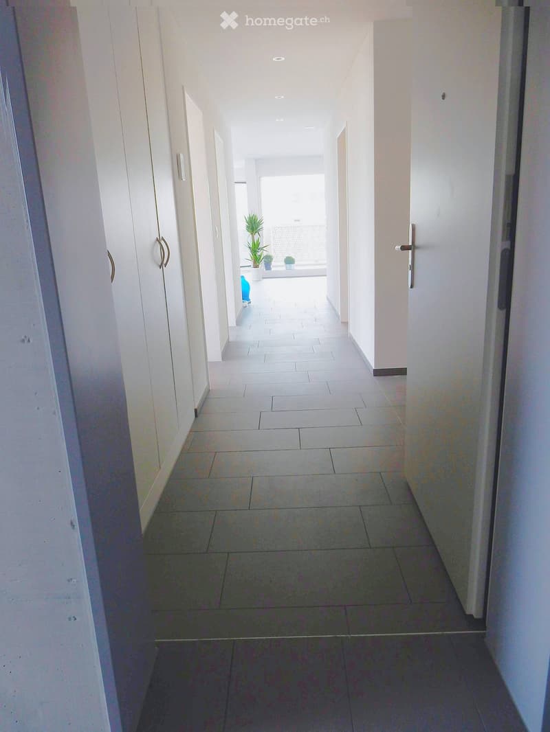 Clean, modern hallway with white walls, tiled flooring, and several doors on both sides. Natural light streams in from the end of the corridor.