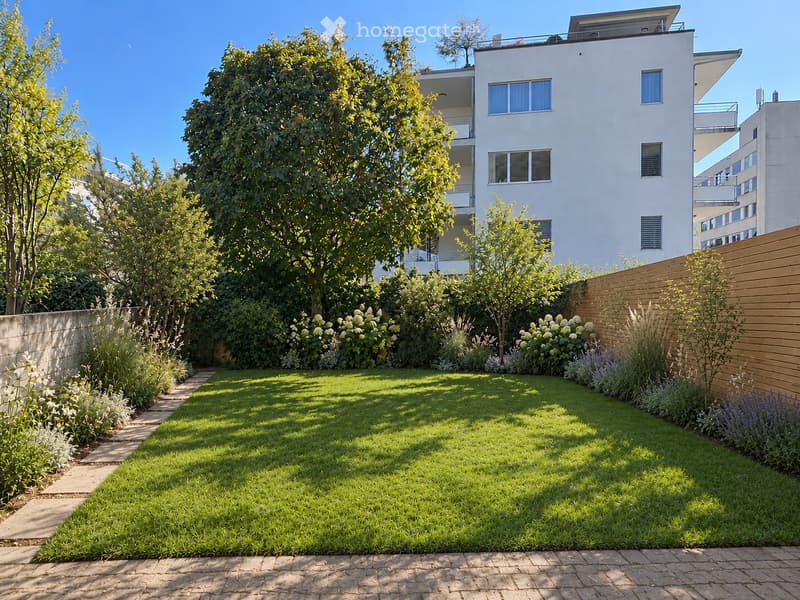 Well-maintained garden with a neatly trimmed lawn, a brick pathway, and various plants and trees, including a wooden fence on one side and a modern building in the background.