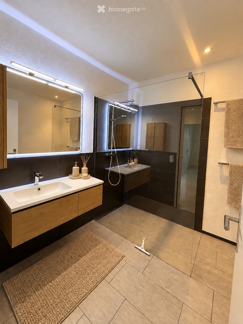 Modern bathroom with double sinks, large mirrors with built-in lighting, a glass-enclosed shower, wooden cabinets, and tiled floors.