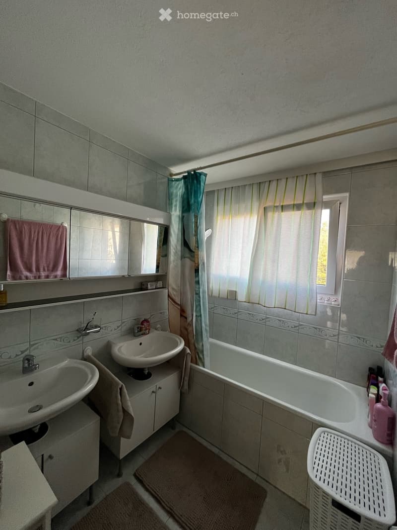 Bathroom with two sinks, a bathtub, a mirror, and a window with a shower curtain. Includes a towel, a bathroom cabinet, and a laundry basket.