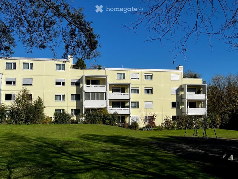 A multi-story residential building with light yellow walls, multiple windows, and balconies, surrounded by a well-maintained lawn and trees.