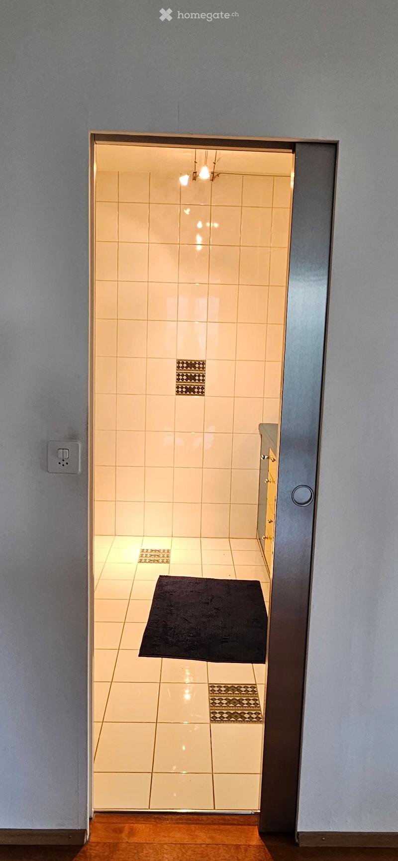 A view of a tiled bathroom entrance from a wooden-floored hallway, featuring a black bath mat on the floor and a glass shower door.