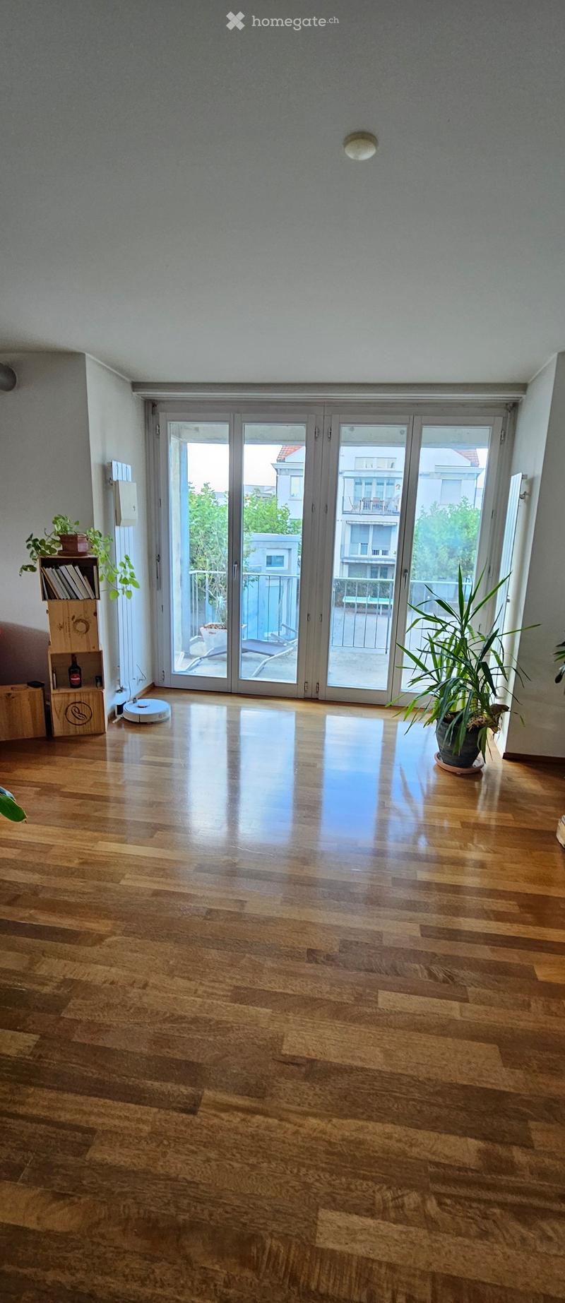 Empty room with wooden flooring, large sliding glass doors leading to a balcony, and a few potted plants near the entrance.
