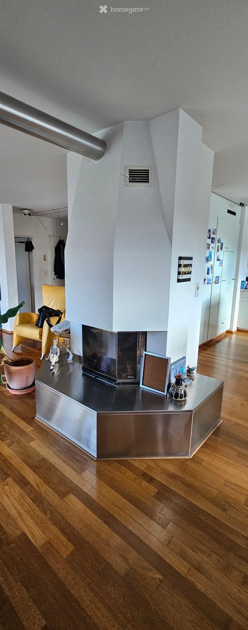 Modern living room with a sleek, metallic fireplace, wooden flooring, a yellow armchair, a small table with decorative items, and a potted plant.