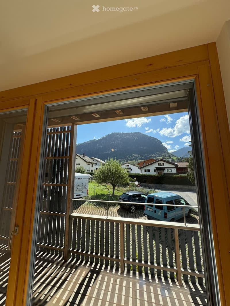 Balcony with wooden railing, overlooking a scenic valley with houses, cars, and mountains in the background.
