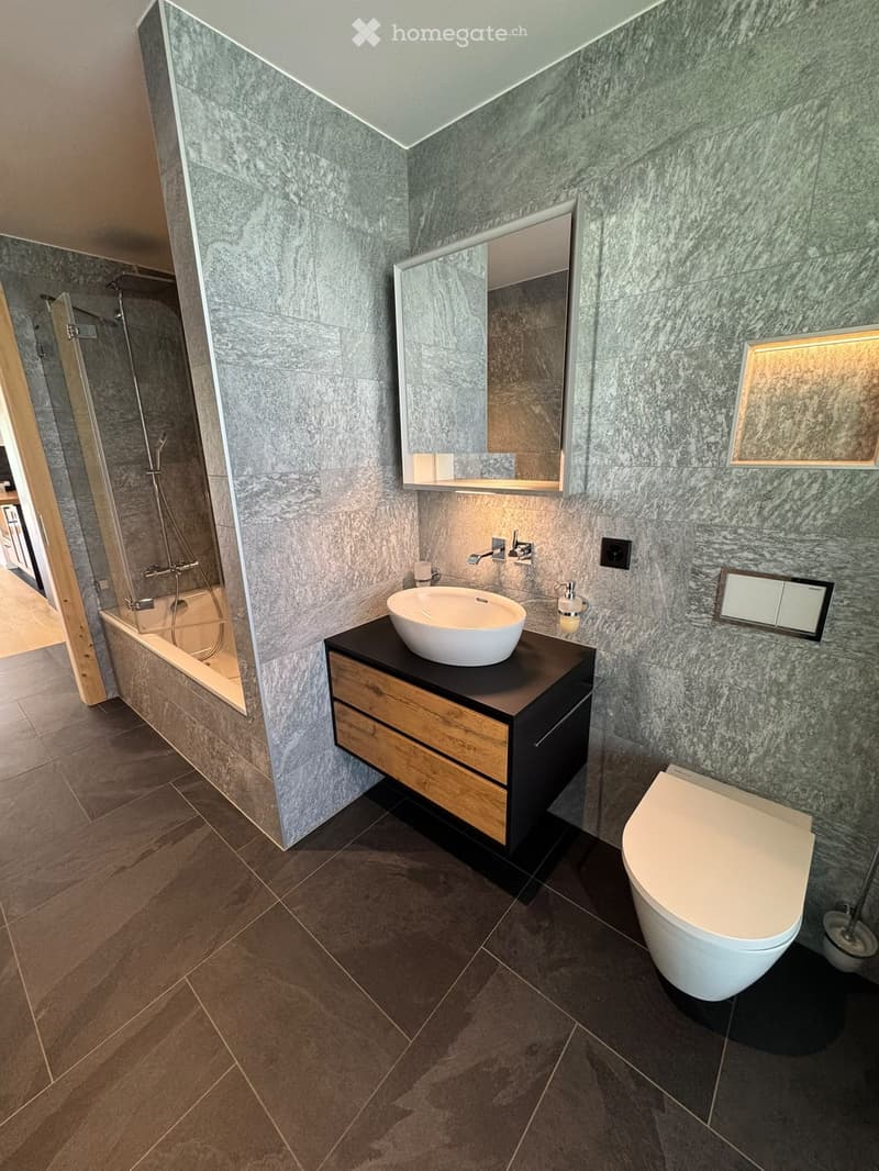 Modern bathroom with grey marble walls, a white sink on a dark wooden vanity, a toilet, a bathtub, and a large mirror above the sink.