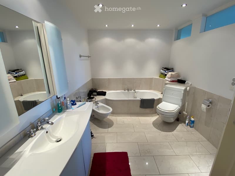 Modern bathroom with a large mirror, double sink, bathtub, toilet, bidet, and neatly stacked towels. Features recessed lighting and a red bathroom rug on the floor.