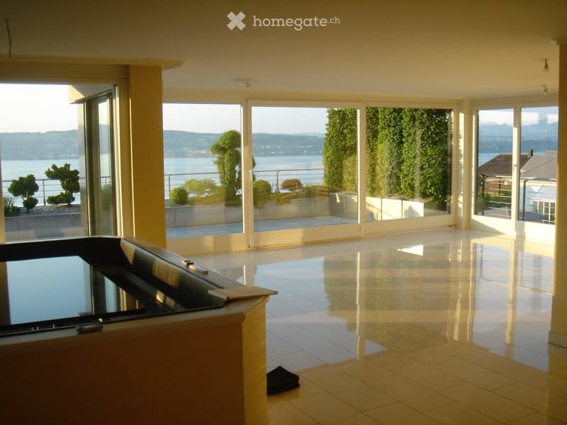 Empty room with large windows overlooking a scenic lake and mountains, polished tile floor, and a small table with a laptop in the foreground.