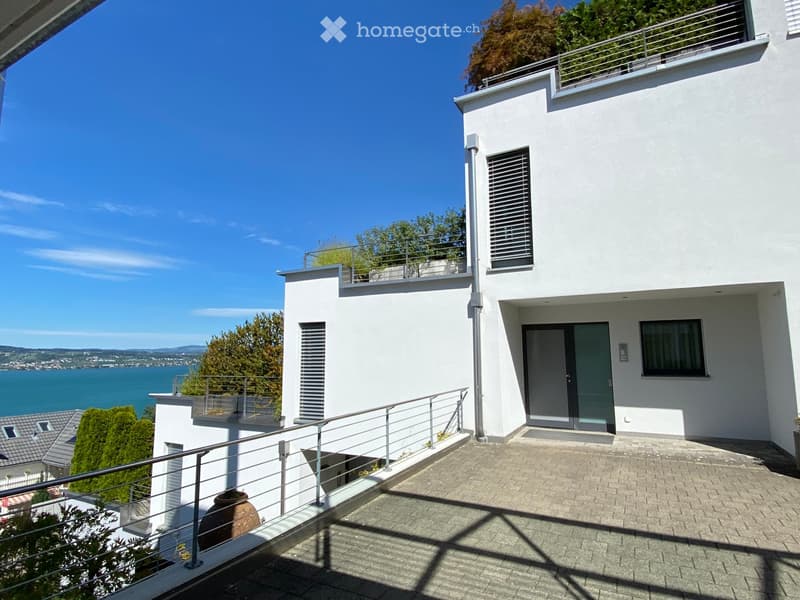 Modern white house with multiple levels, balconies, and a scenic view of a lake and distant mountains.