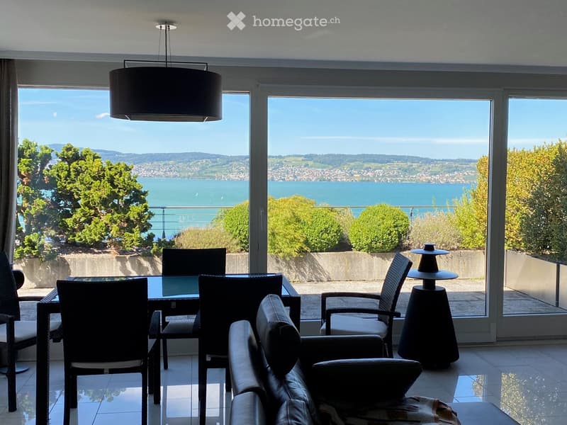 Modern living room with large windows offering a scenic view of a lake and surrounding landscape. Features a sofa, chairs, a table, and a lamp.