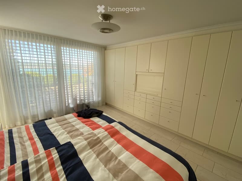 Bedroom with a large bed covered in a red, white, and blue checkered duvet, a wardrobe with multiple drawers, and a window with sheer curtains.