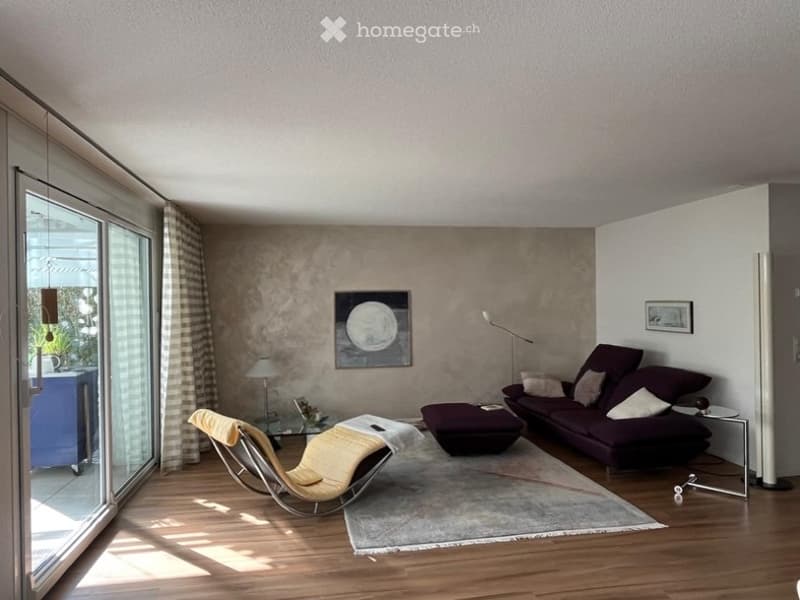 Modern living room with large windows, wooden floor, grey sofa, ottoman, yellow chair, small table, and artwork on the wall.