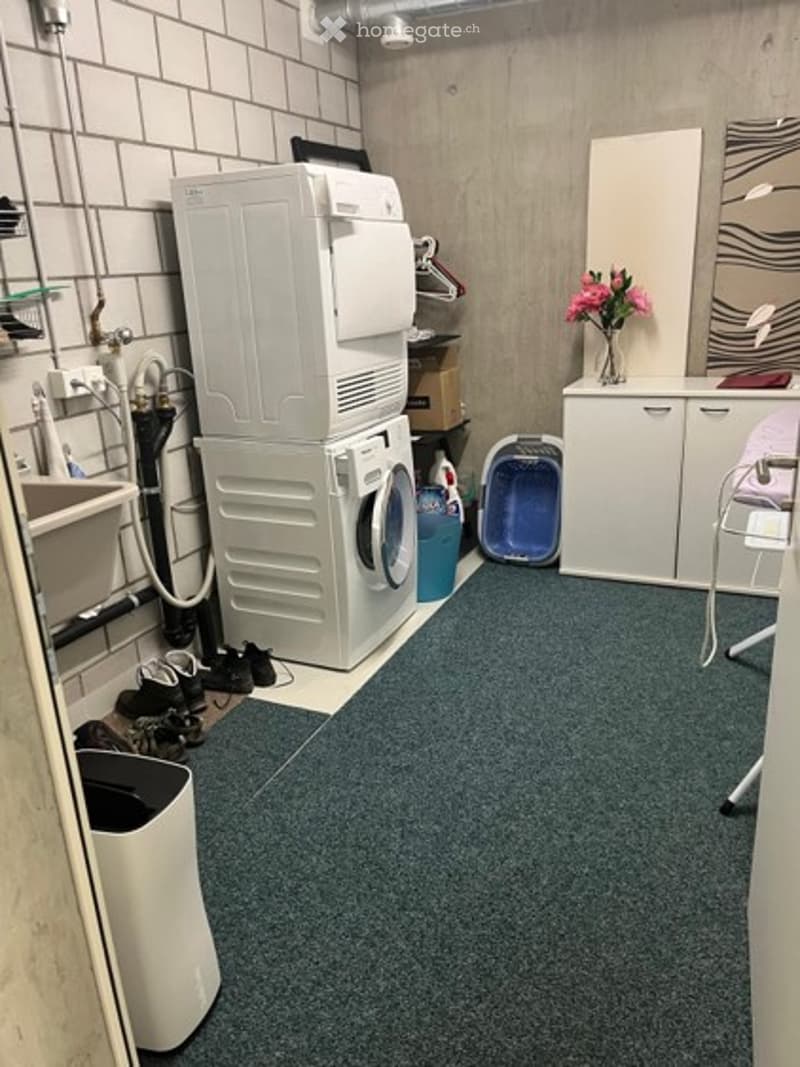 A small, functional laundry area with a washing machine, dryer, sink, and cabinet. There is a vase with pink flowers on the cabinet and a small stool nearby.