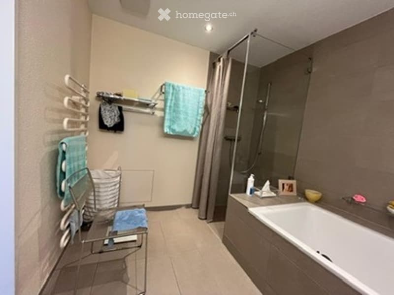 Modern bathroom with a bathtub, towel rack, shower, and small stool. Towels and a picture frame are visible on the counter.