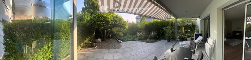 A modern outdoor patio area with a glass-enclosed balcony overlooking a lush garden. The patio features a round table with chairs, lounge chairs, and a striped awning providing shade. The garden includes trees, shrubs, and decorative elements.