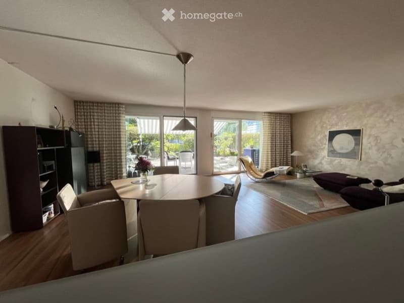 Bright, open-plan living and dining area with wooden flooring, large windows, and a view of the garden. Features a round dining table with chairs, a sofa, a lounge chair, and a bookshelf.