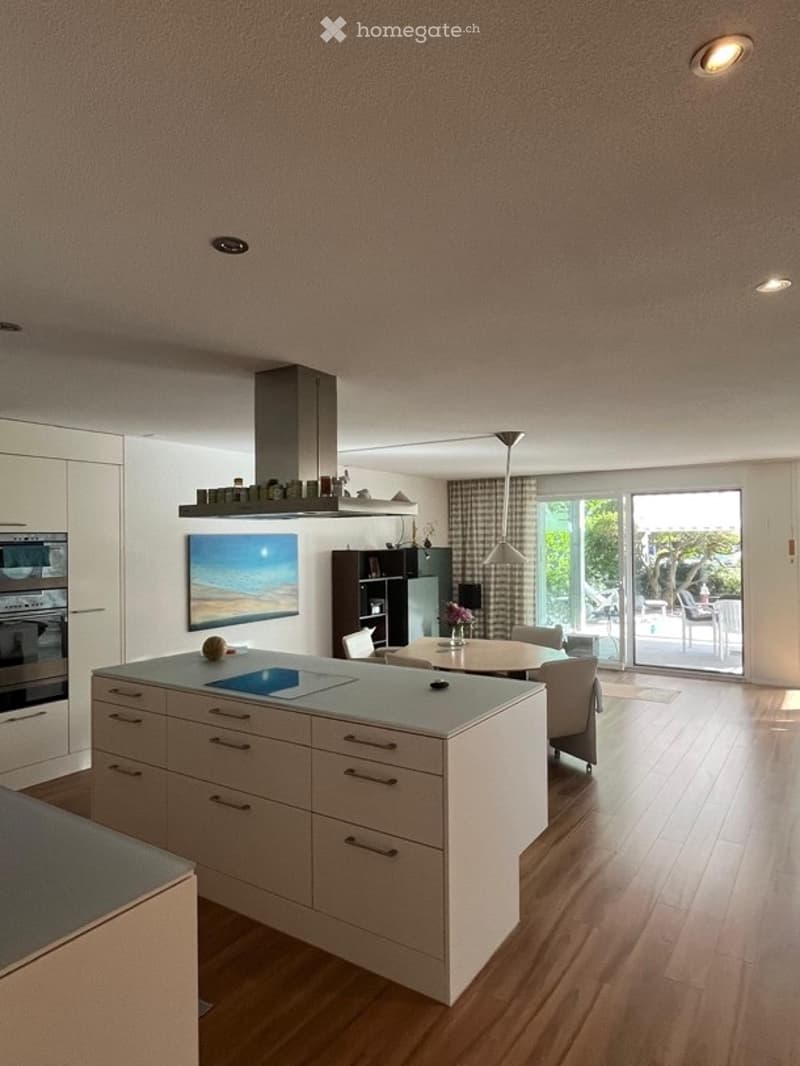 Modern kitchen with white cabinets, a central island, built-in stove and oven, and a dining area with chairs and a table. Large windows and a sliding glass door lead to a balcony or terrace area.