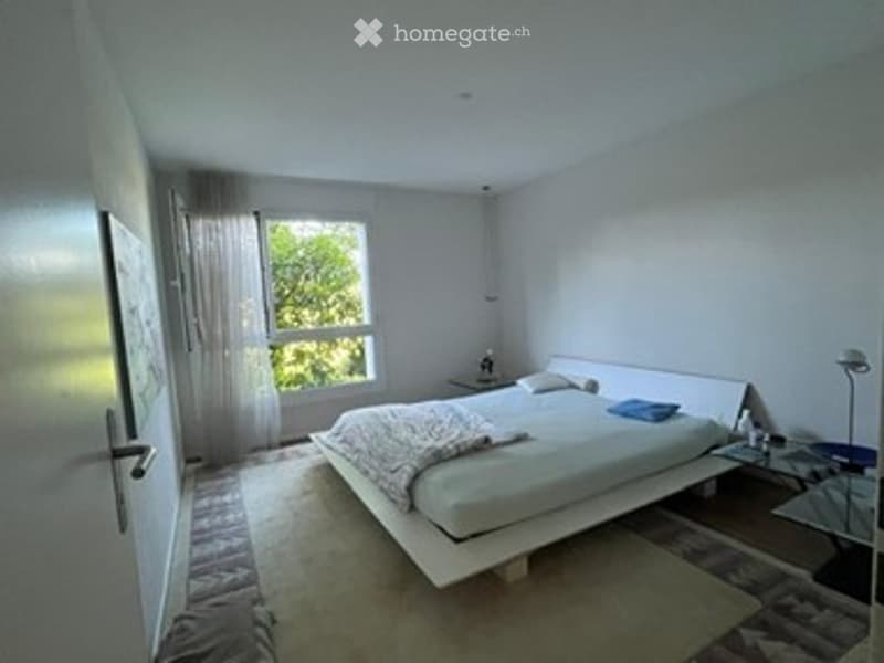 Modern, minimalist bedroom with a white bed, nightstand, lamp, and large window with sheer curtains.