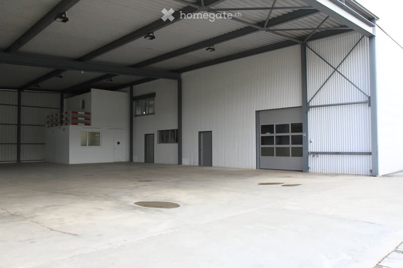 Large industrial building with corrugated metal walls, multiple doors, and windows. The interior shows a spacious, open concrete floor with visible structural beams and lighting fixtures.