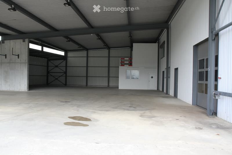 Large, empty industrial-style warehouse with concrete floors, metal walls, and large roll-up doors. Minimal lighting and no furniture or equipment present.
