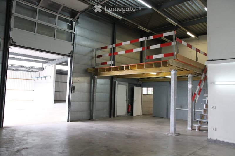 Empty industrial-style garage with concrete floors, metal walls, large windows, and a wooden loft area with safety barriers.