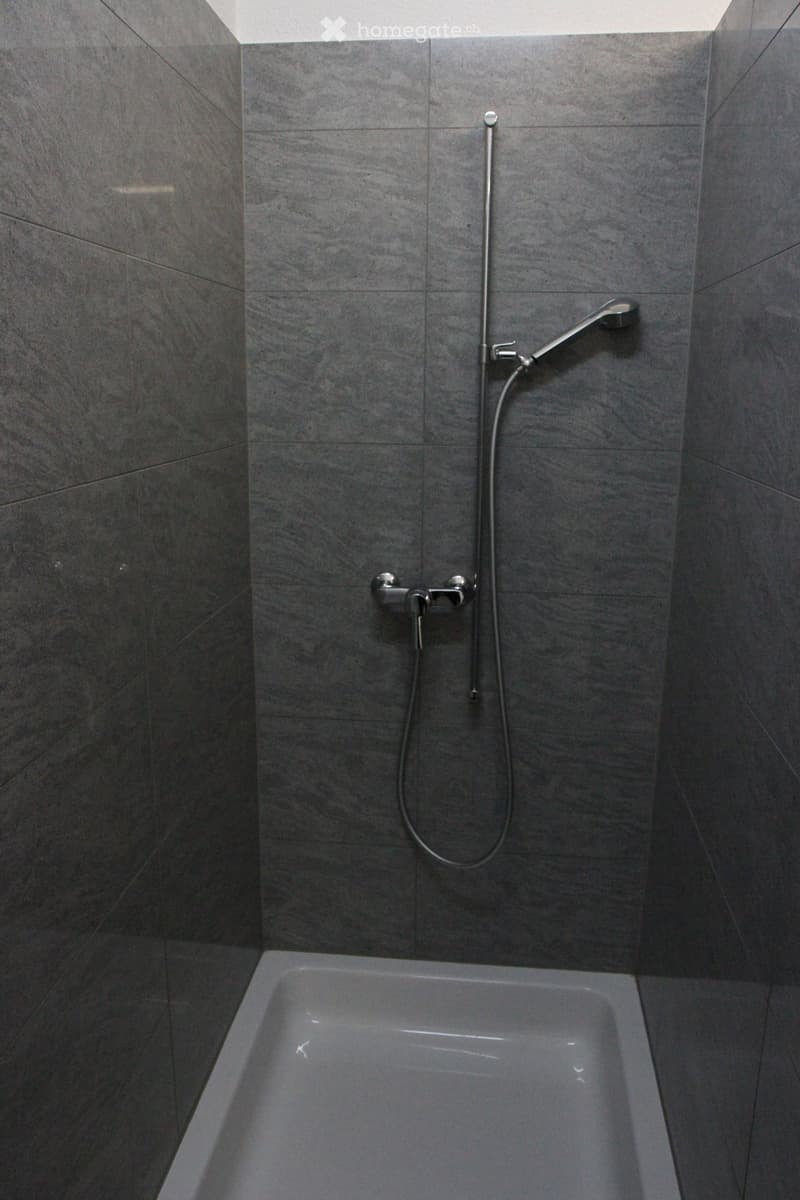 Modern bathroom with a bathtub, grey tiled walls, and a handheld showerhead with a hose.