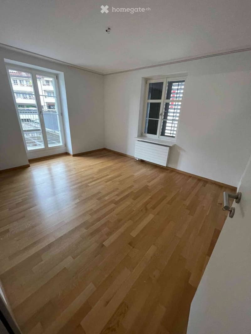 Empty room with wooden flooring, white walls, and large windows, including a radiator and a door handle visible.