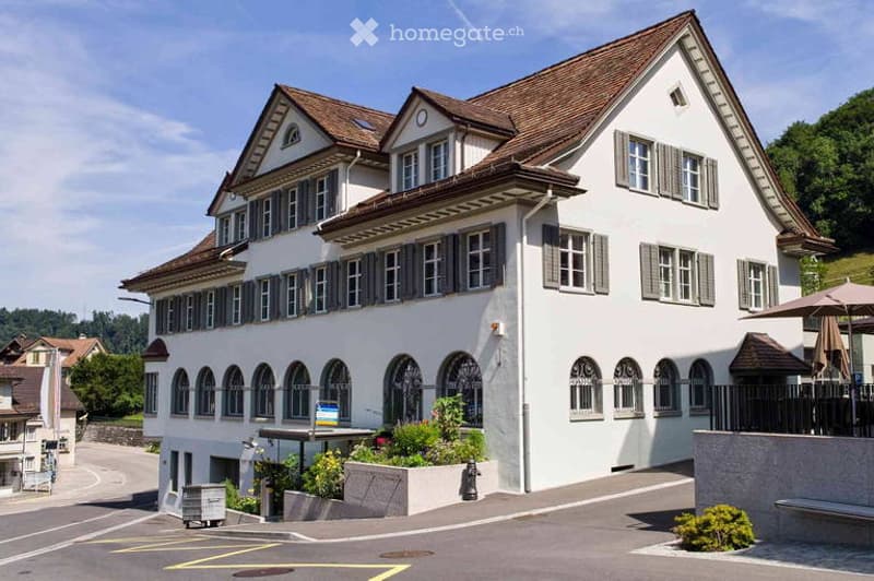 A large, multi-story white building with a brown roof and multiple windows, featuring a well-maintained garden area in front.