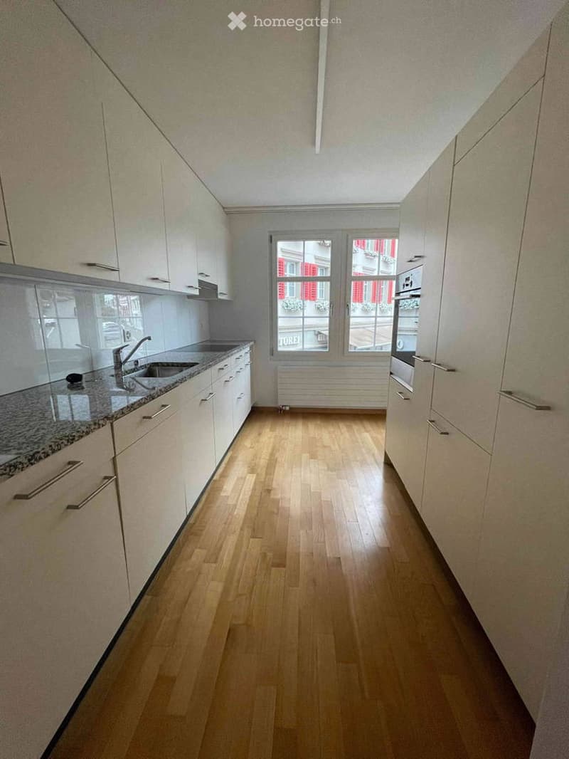 Empty kitchen with light-colored cabinets, granite countertop, wooden floor, and large windows showing a view of a building with red shutters.