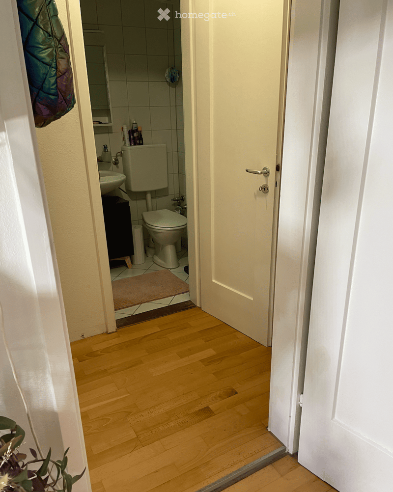 Bathroom with white tiled walls, a toilet, sink, mirror, and a small cabinet. The floor is tiled, and a brown rug is placed in front of the toilet.
