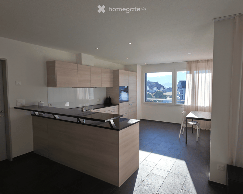 Modern kitchen with light wood cabinets, black countertop, and a small dining area with a table and chairs. Large windows provide natural light and a scenic view.