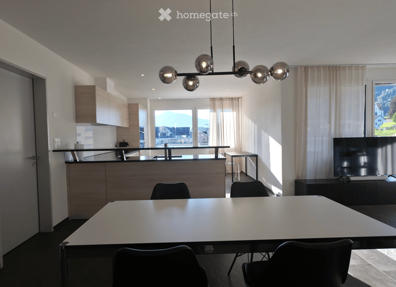Open-plan living and dining area with a large table, chairs, kitchen island, and television. Natural light streams in from large windows with curtains.