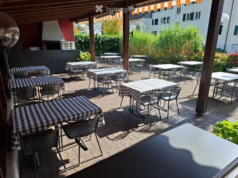Outdoor seating area with multiple tables and chairs covered with checkered tablecloths, surrounded by greenery and a building in the background.