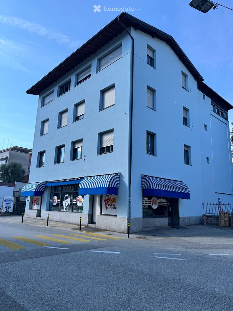 Two-story commercial building with light blue facade, multiple windows with shutters, striped awnings, and signage for "POMS market suisse".