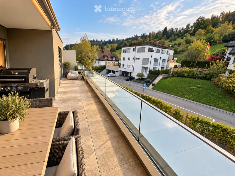 Balcony with a wooden table, wicker chairs, a sofa, a grill, and potted plants, overlooking a scenic landscape with houses and greenery.