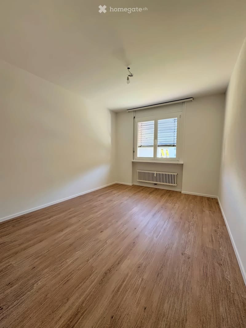 Empty room with wooden flooring, white walls, a window with blinds, and a radiator below the window.