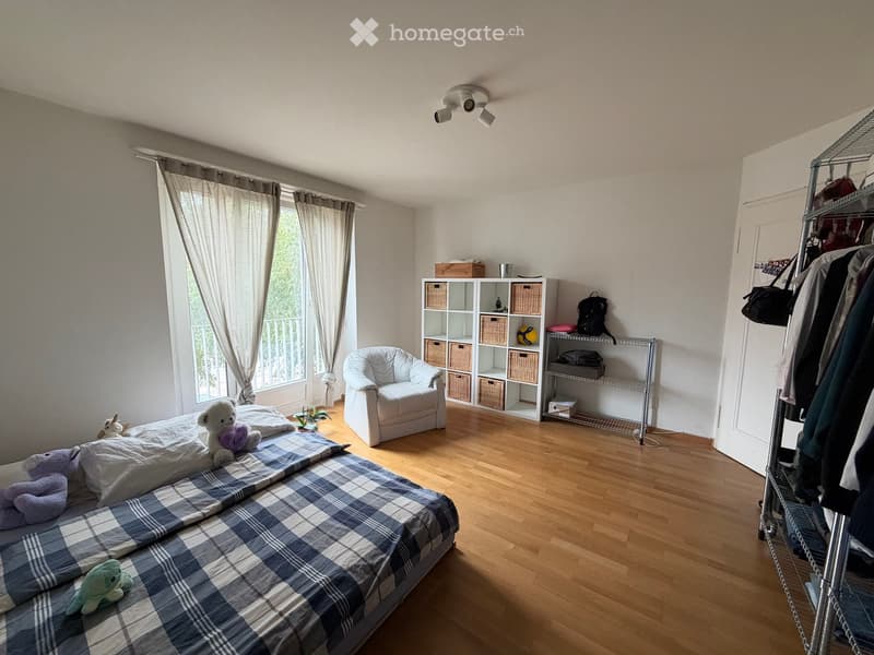 Bright bedroom with wooden flooring, large window with curtains, bed with plaid blanket, stuffed animals, armchair, and storage unit with baskets.