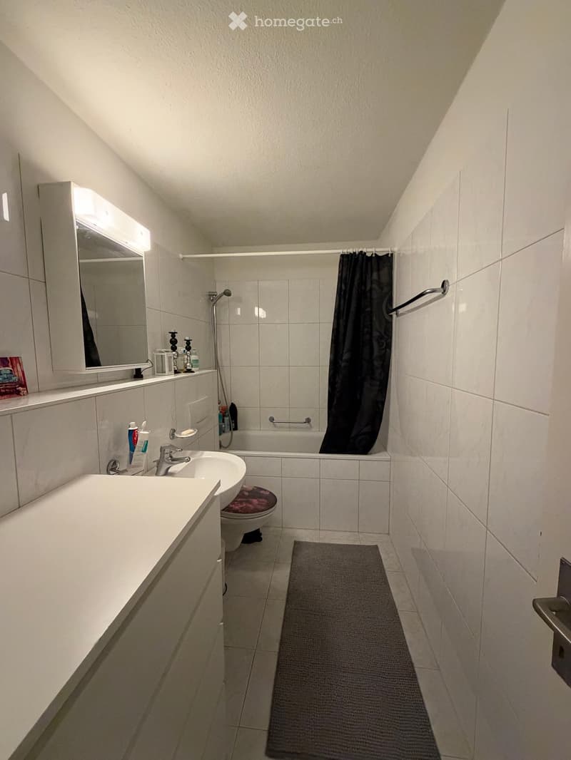 Modern bathroom with white tiles, a sink with a faucet, a mirror with a built-in cabinet, a bathtub with a shower curtain, a toilet, and a towel rack. The space includes a gray bathroom rug and various toiletries on the sink counter.