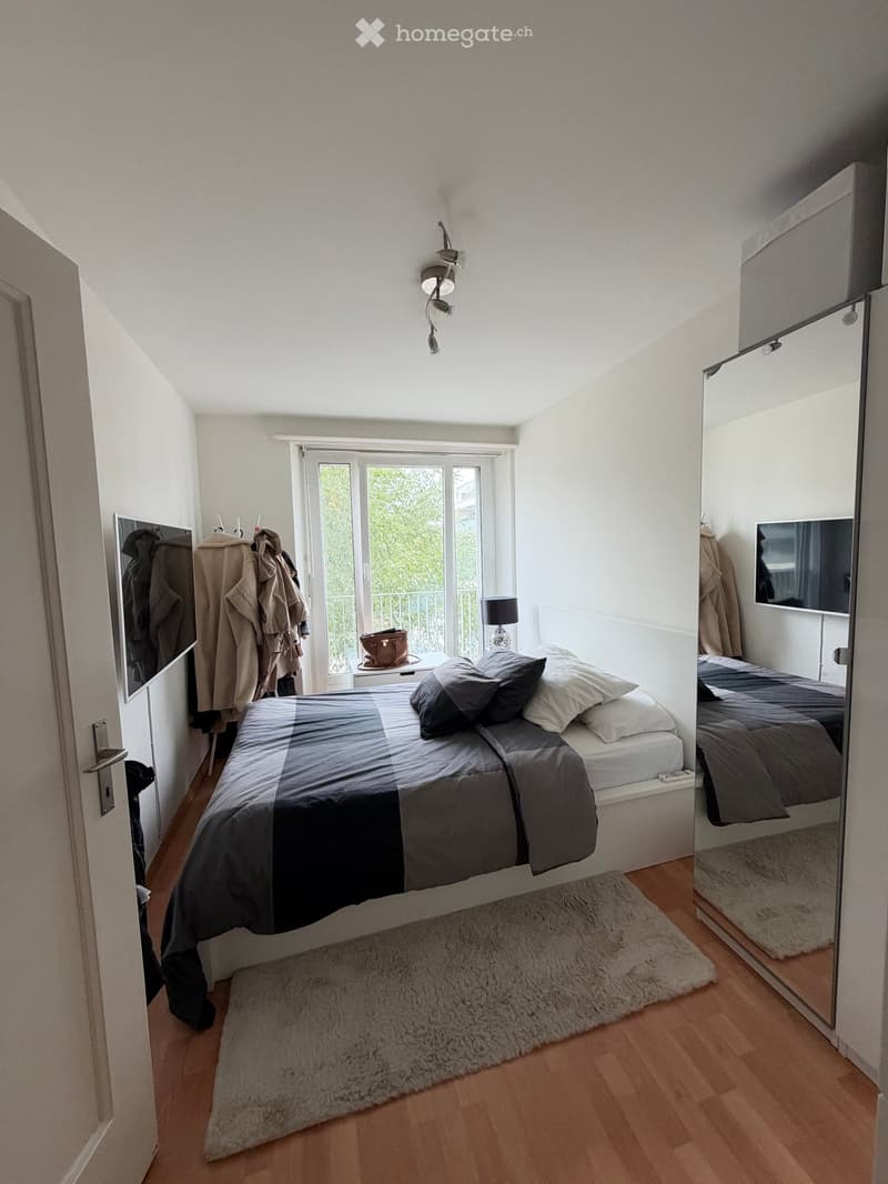 Modern bedroom with a large bed, grey bedding, a fluffy rug, a wall-mounted mirror, a small table with a lamp, and coats hanging on the wall.