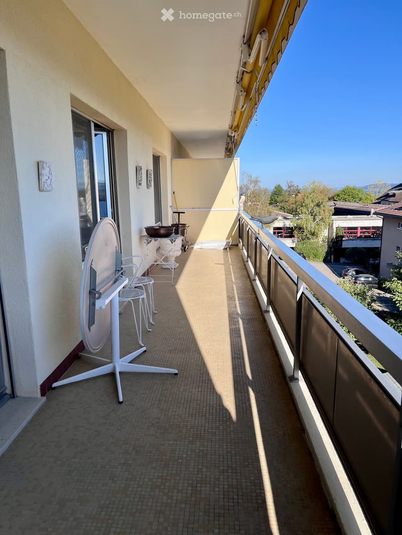 Balcony with tiled floor, white metal chair, small table, and umbrella stand, overlooking a residential area with trees and buildings.