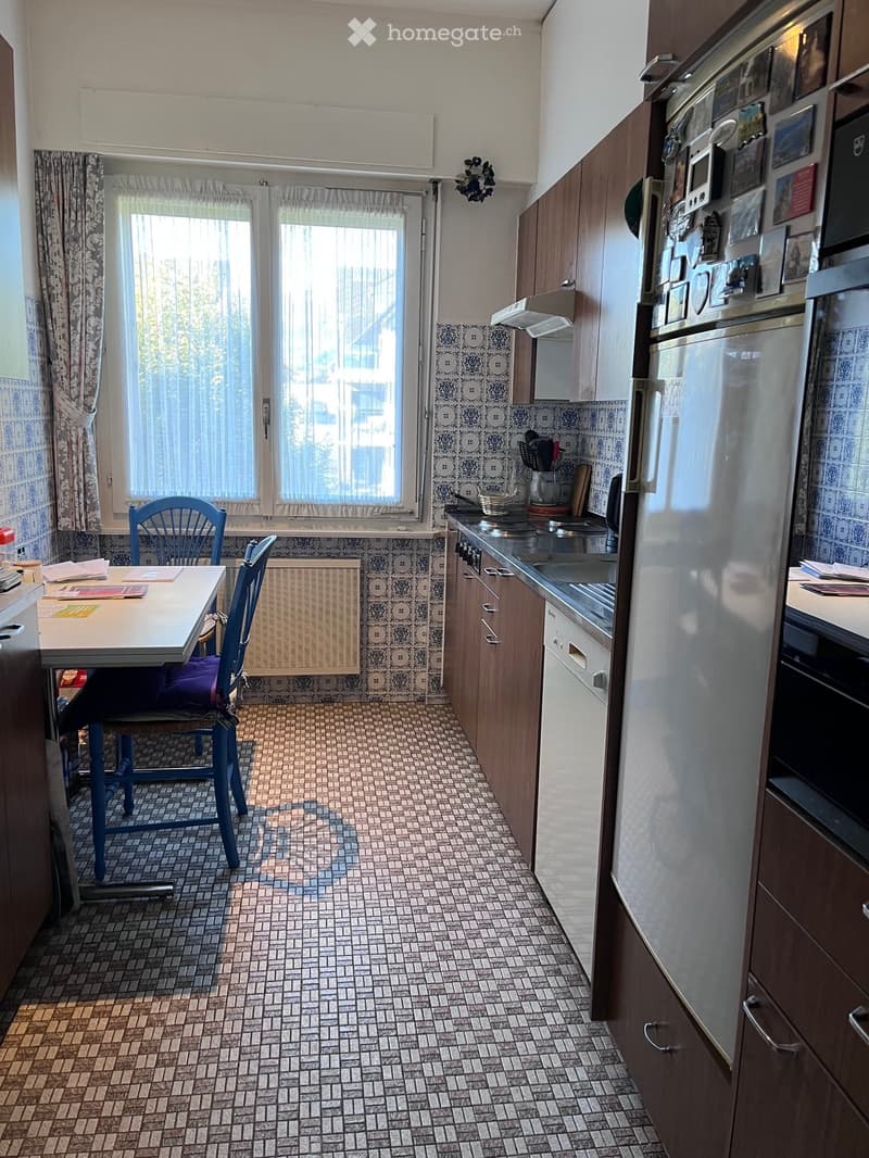 A compact kitchen with a dining area, featuring a tiled floor, wooden cabinets, a refrigerator, and a small table with chairs.