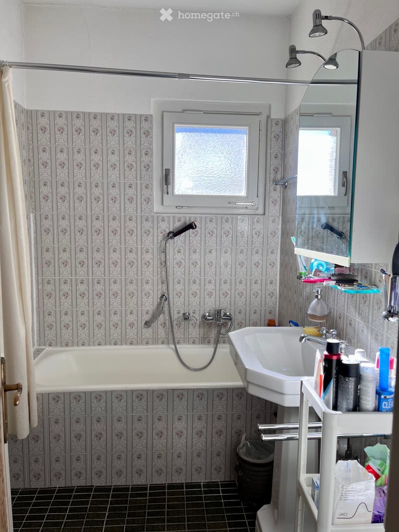 Bathroom with a bathtub, sink, mirror, and various toiletries. The walls are tiled with a floral pattern, and there is a window above the sink.