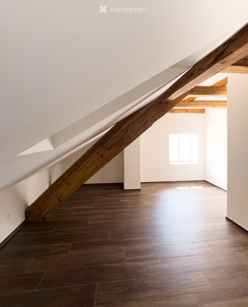 Empty attic space with wooden floor, white walls, exposed wooden beams, and a small window letting in natural light.