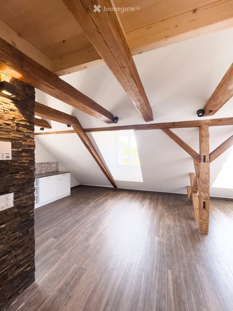 An empty attic space with exposed wooden beams, wooden flooring, and a stone accent wall. Natural light streams in through a skylight window.