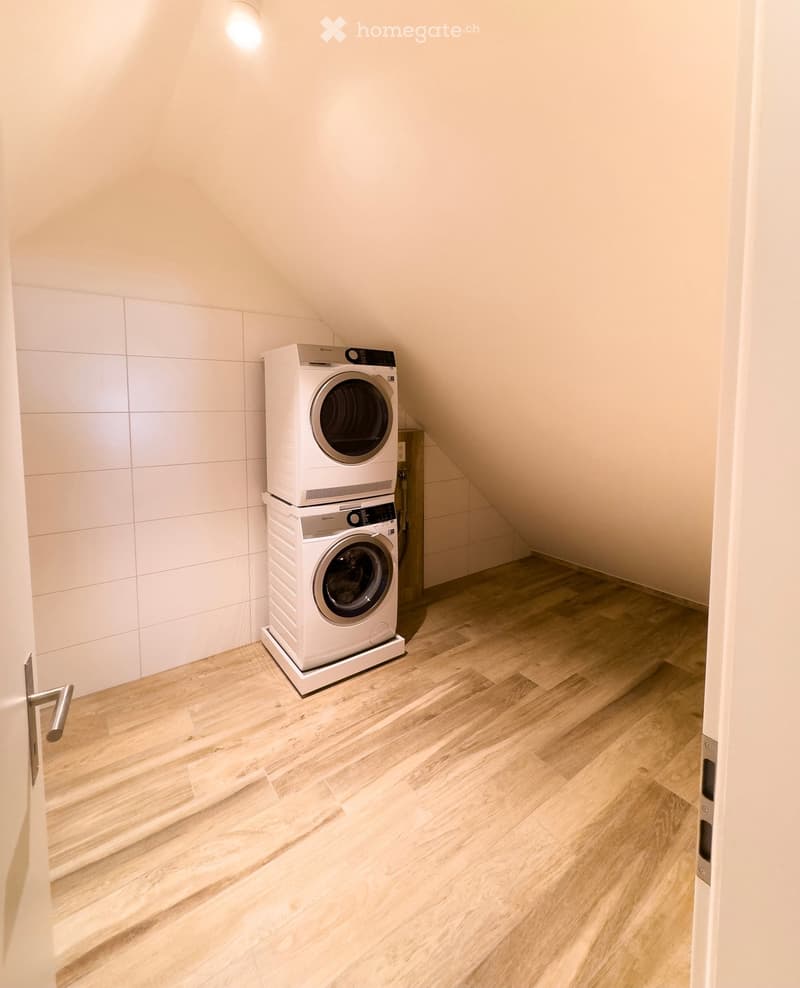 Modern laundry room with light wooden flooring, white tiled walls, and stacked washing machine and dryer.
