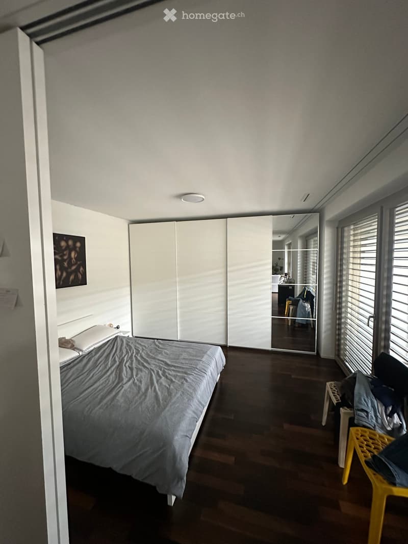 Modern bedroom with a large bed, dark wooden flooring, white wardrobe, and a yellow plastic chair near the window.