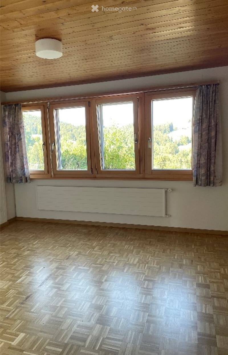 Empty room with wooden flooring, large windows with wooden frames, and a radiator below the windows. The ceiling is wooden with a central light fixture.