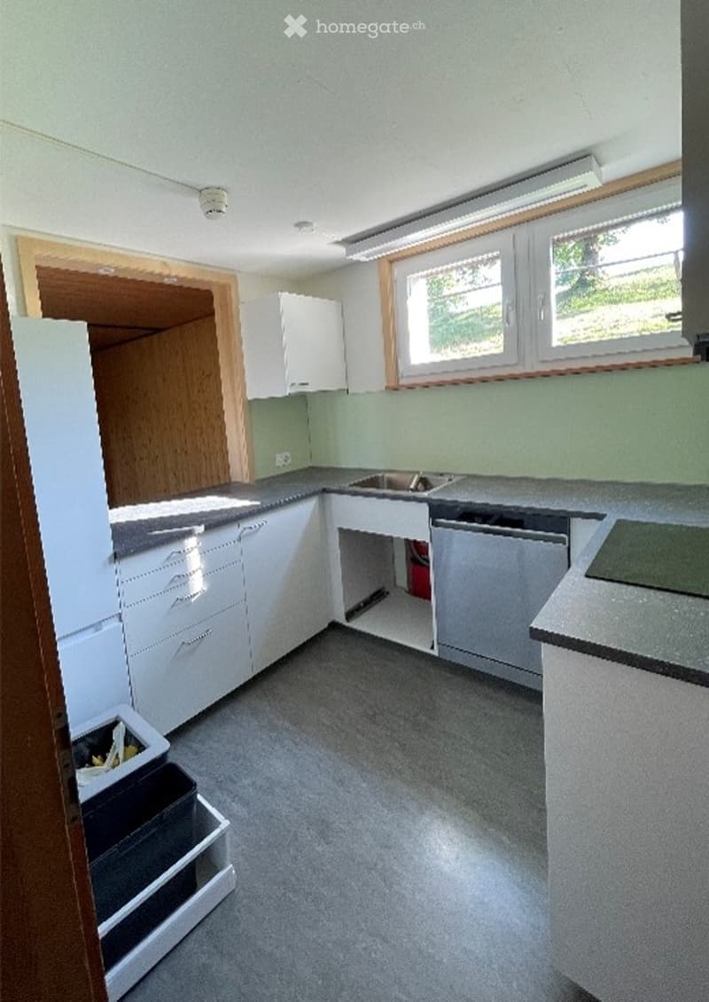 Empty kitchen with white cabinets, gray countertops, and a sink. The space includes a refrigerator and a partially open cabinet, with natural light coming through windows.