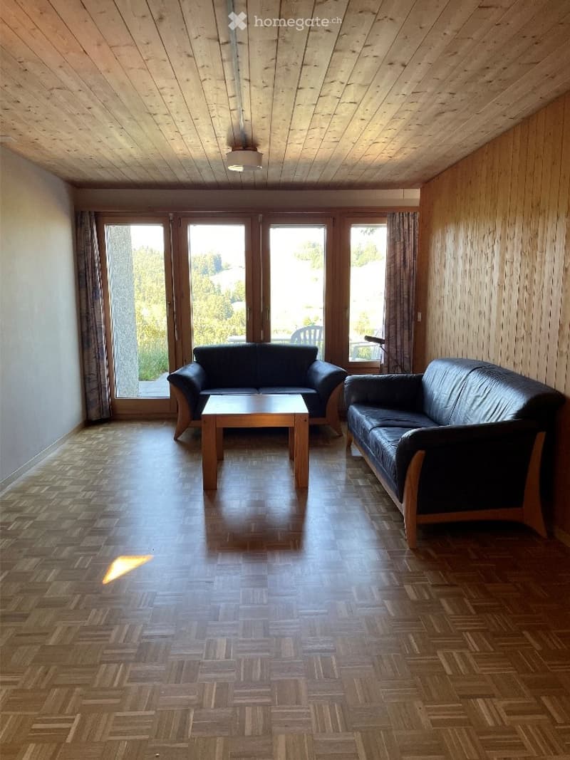 Bright living room with wooden parquet flooring, two black leather sofas, a wooden coffee table, large windows with curtains, and a view of the outdoors.