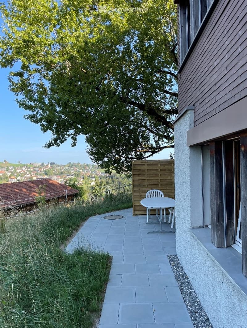 Outdoor patio area with a small table and chairs, surrounded by greenery and a wooden fence, offering a scenic view of the town and landscape.